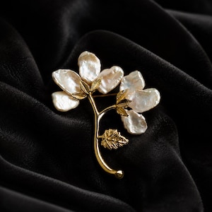May include: A gold-toned metal brooch shaped like a flowering branch, featuring multiple irregular white baroque pearls as petals and small textured gold leaves attached to the stem, creating a botanical design with a polished metallic finish.