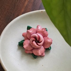May include: A floral brooch featuring layered, light pink petals with a glossy finish. The center of the flower is a darker pink, and small, pointed green leaves are visible tucked between the outer petals of the rose-shaped accessory.