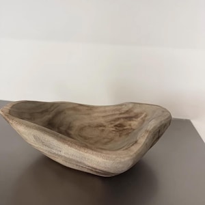 May include: A wooden bowl with a natural, light brown finish. The bowl has an organic, asymmetrical shape with a smooth interior and textured exterior. The wood grain is visible, adding to its rustic charm. This decorative bowl is suitable for home decor.