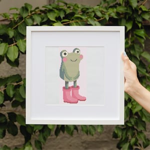 May include: A framed cross-stitch art piece featuring a smiling frog wearing pink rain boots. The frog is light green with rosy cheeks and large eyes. The background is a light pink vertical stripe.