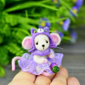 May include: A handmade miniature white mouse wearing a purple cat-eared hat and a matching lace dress. The mouse has pink ears and a pink tail. It is holding a green and purple bead. The background is blurred green foliage.