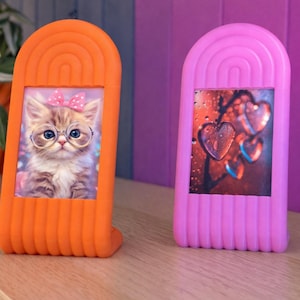 Puede incluir: Dos marcos de fotos decorativos. Uno es naranja y presenta una imagen de un gatito con gafas y un lazo. El otro es rosa y muestra una imagen de formas de corazón. Ambos marcos tienen un diseño superior arqueado único.