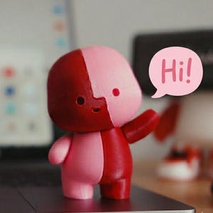 Peut inclure: Figurine bicolore rose et rouge avec une bulle de dialogue disant "Hi!". La figurine est posée sur un ordinateur portable. L'arrière-plan comprend un écran d'ordinateur portable flou et d'autres objets.