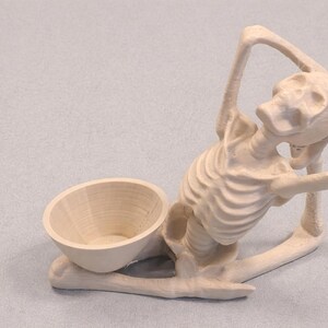 May include: A light-colored wooden skeleton figurine with a bowl-shaped receptacle. The skeleton is posed in a seated position, with arms raised. The bowl is positioned near the skeleton's legs. The figurine is made of wood.