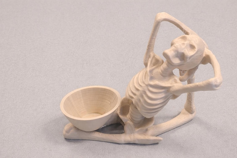 May include: A light-colored wooden skeleton figurine with a bowl-shaped receptacle. The skeleton is posed in a seated position, with arms raised. The bowl is positioned near the skeleton's legs. The figurine is made of wood.