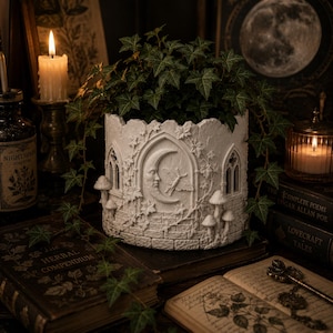 May include: A white gothic planter, adorned with a crescent moon and moth motif, overflowing with green ivy. The planter has architectural features, including arched windows and mushroom details. The scene is set with candles and antique books.
