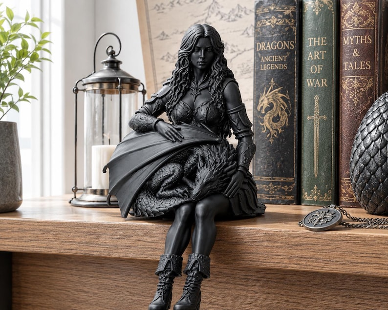 May include: A matte black figurine of a long-haired warrior in armor sitting down while cradling a small dragon with folded wings. The figure wears boots and textured armor, with the dragon resting on their lap. The sculpture is a solid dark color.