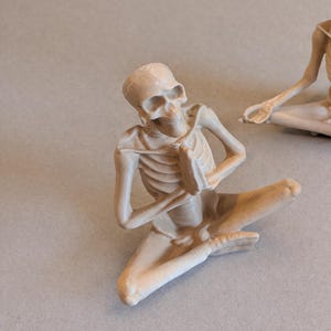 May include: A beige-colored skeleton figurine in a seated yoga pose with hands together in prayer. The figurine is detailed with visible ribs and skull features. Another similar figurine is partially visible in the background.