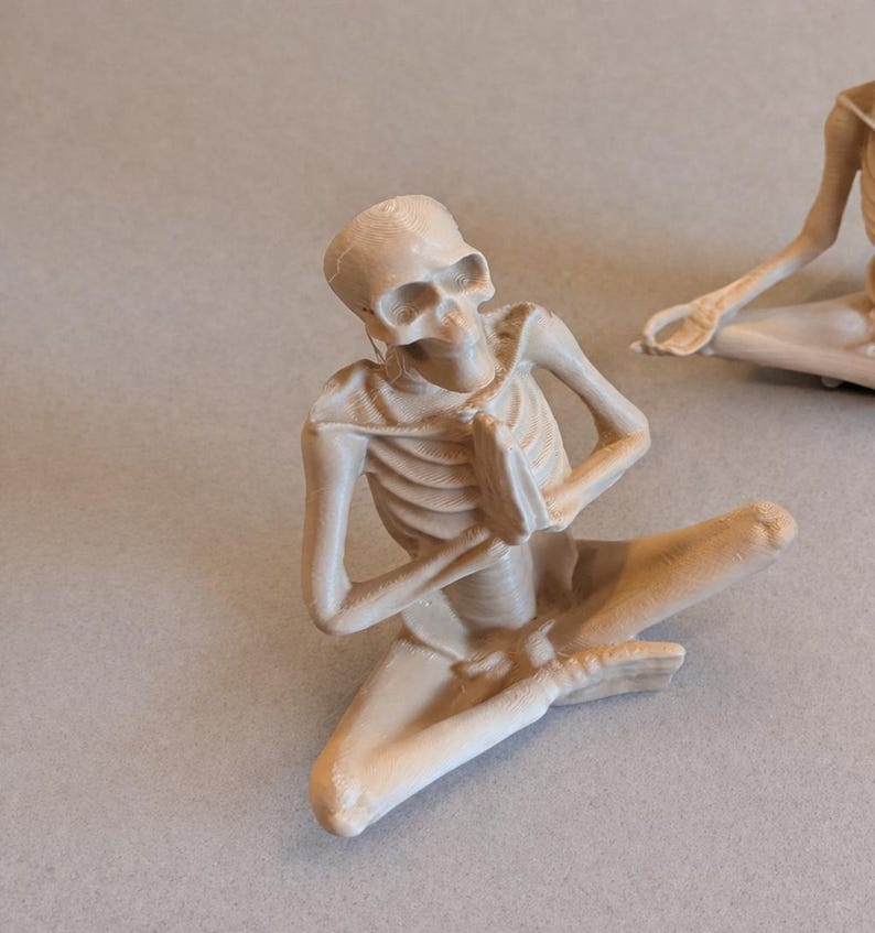 May include: A beige-colored skeleton figurine in a seated yoga pose with hands together in prayer. The figurine is detailed with visible ribs and skull features. Another similar figurine is partially visible in the background.