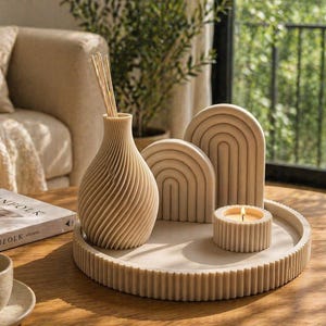 Könnte beinhalten: Ein beiges Dekorationsset auf einem runden Tablett. Das Set beinhaltet eine Vase mit Schilf, zwei bogenförmige Skulpturen und einen kleinen Kerzenhalter mit einer brennenden Kerze. Das Tablett und die Objekte haben ein geripptes Design. Ein Buch und eine Tasse sind ebenfalls sichtbar.