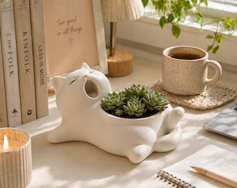 May include: A white ceramic cat-shaped planter, yawning with a succulent arrangement inside. The planter is on a desk with books, a candle, a coffee mug, and a framed print that says "See good in all things".