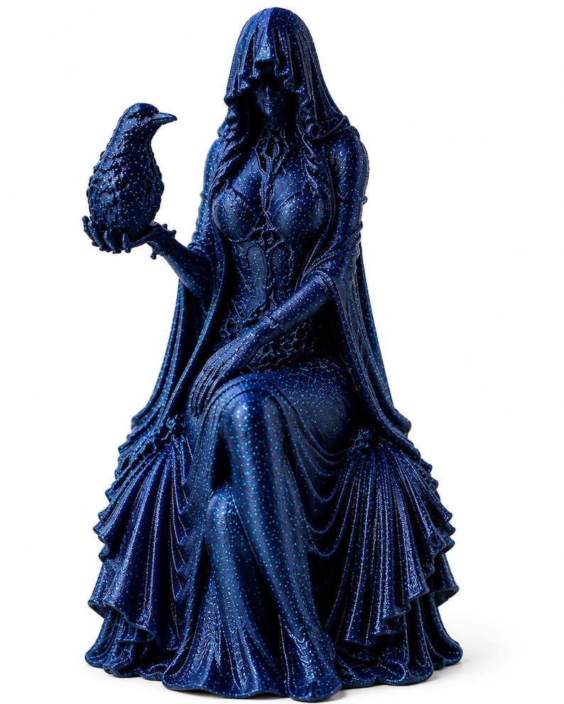 May include: A dark blue glittery figurine of a seated hooded figure wearing a long flowing gown with intricate bodice detailing. The figure holds a small bird perched on one hand, with the hood obscuring the face and the gown draped in folds.
