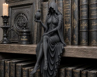 May include: A matte black figurine of a hooded figure sitting with legs crossed, holding a small raven in one hand. The figure wears a long, draped gown with intricate lace-up detailing on the bodice and sleeves, and a delicate anklet on one foot.