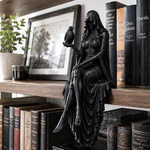 May include: A black figurine of a hooded figure sitting on a shelf edge, wearing a long flowing gown and holding a small bird in one hand. The figure has detailed textures on the bodice and skirt, with bare feet hanging down over the shelf edge.