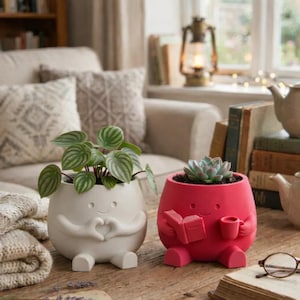 May include: Two whimsical, smiling ceramic planters. One is white with arms forming a heart shape, holding a green plant. The other is red, holding a book and a mug, with a succulent inside. Both have small feet.
