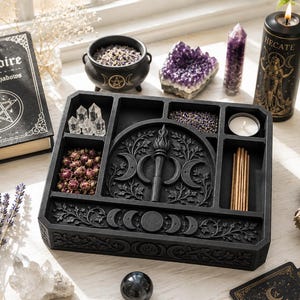 May include: A black wooden altar tray featuring a central relief of a torch and triple moon symbol, surrounded by compartments containing clear quartz points, dried rosebuds, lavender, and incense sticks, with moon phase carvings on the front edge.