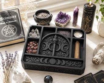 Puede incluir: Una bandeja de altar de madera negra con un relieve central de una antorcha y el símbolo de la triple luna, rodeada de compartimentos con puntas de cuarzo, capullos de rosa secos, lavanda e incienso, con tallas de fases lunares.