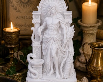 May include: A white resin statue of the god Apollo standing between two columns, featuring a sunburst halo, a lyre, a snake coiled around a pedestal, a quiver of arrows, and a laurel branch, all set on a rectangular base with floral carvings.