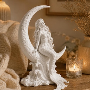 Puede incluir: Una escultura de sirena blanca sentada en una luna creciente, sobre olas estilizadas. La sirena tiene cabello largo y suelto y lleva joyas delicadas. Es un objeto decorativo para la decoraci&oacute;n del hogar.