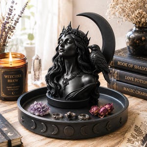 May include: A black sculpture of a woman with a crown and raven on a crescent moon, displayed on a tray with rings, crystals, and dried flowers. A candle with the text "Witches Brew" is also visible. The scene is set on a wooden surface with books.