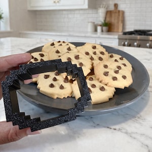 Könnte beinhalten: Ein Teller mit Schokoladen-Chip-Cookies und einem schwarzen, pixeligen Ausstecher. Die Kekse sind goldbraun mit Schokoladenstückchen. Der Ausstecher wird in einer Hand gehalten.