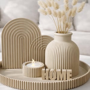 Peut inclure: Un plateau décoratif beige avec un vase contenant des fleurs séchées, deux sculptures en forme d'arche, un bougeoir avec une bougie allumée et le mot "HOME". Les objets sont disposés sur une table blanche.