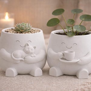 Könnte beinhalten: Zwei weiße Keramik-Pflanzgefäße mit lächelnden Gesichtern. Eines hält eine schlafende Katze, das andere eine Teetasse. Ein Topf hat eine Sukkulente, der andere eine grüne Pflanze. Die Töpfe stehen auf einer neutralfarbenen Oberfläche.