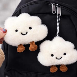 May include: Two fluffy white cloud-shaped keychains with smiling faces and brown feet. One keychain is attached to a black backpack with a silver chain. The other cloud is held by a hand. The keychains are made of soft, plush material.