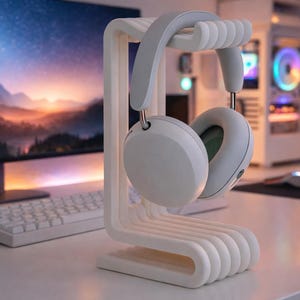 May include: White headphones resting on a matching white headphone stand. The stand has a curved design with multiple horizontal bars. The headphones are light gray and white, with a minimalist aesthetic. A computer monitor, keyboard, and mouse are in the background.