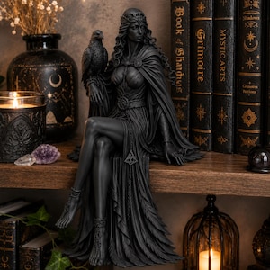 May include: A matte black figurine of a seated figure wearing a crown and a long, flowing gown with feather-like textures. The figure has a bird perched on one arm and wears intricate anklets. The sculpture is crafted with detailed features.