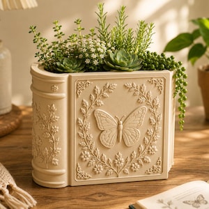May include: A cream-colored rectangular planter shaped like a book with a relief butterfly design surrounded by floral vines on the front panel. The planter is filled with various green succulents and small leafy plants, creating a textured display.