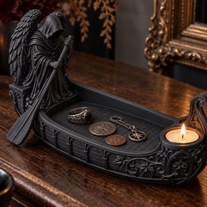 May include: A black resin boat-shaped trinket tray featuring a hooded figure with wings holding an oar at the stern. The interior holds a ring, two coins, and a pendant. A small circular tea light candle holder is integrated into the bow.