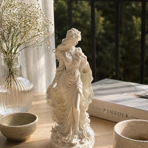 Puede incluir: Una figura blanca de una persona de pie con el cabello largo y fluido y ropa drapeada, colocada sobre una base circular. La escultura presenta detalles intrincados en los pliegues de la tela y el cabello, capturando una pose elegante.