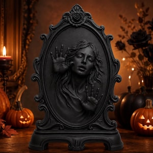 May include: A black oval-shaped decorative plaque featuring a high-relief sculpture of a person with eyes closed, pressing both hands against the surface. The frame is ornate with scrollwork and a rose at the top, set on a sturdy base.