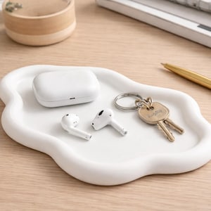 May include: A white, cloud-shaped tray holds a white charging case, two white earbuds, and a set of keys. A gold pen and a keyboard are in the background. The tray is on a light wood surface.
