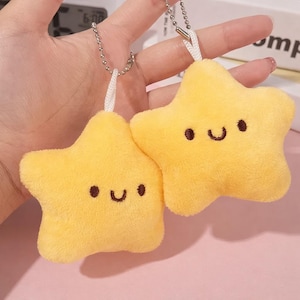 May include: Two yellow star-shaped plush keyrings with smiling faces. Each star has small black dots for eyes and a curved black line for a mouth. The keyrings are attached to silver ball chains with white loops.