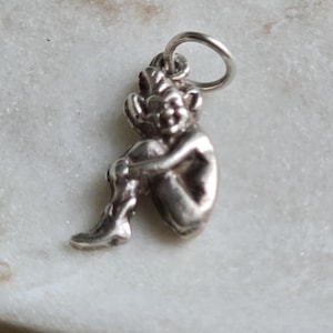 Puede incluir: Un pequeño dije plateado con la forma de un duende o goblin sentado. El dije tiene detalles, incluyendo una cara sonriente, orejas puntiagudas y piernas cruzadas. Está unido a un pequeño anillo para facilitar su fijación.