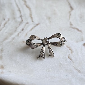 Op de afbeelding: Zilverkleurige strikbroche met heldere, ronde edelstenen. De broche heeft de vorm van een strik, met een centrale edelsteen en kleinere stenen die de lussen van de strik omlijnen. Geplaatst op een wit en grijs marmeren oppervlak.