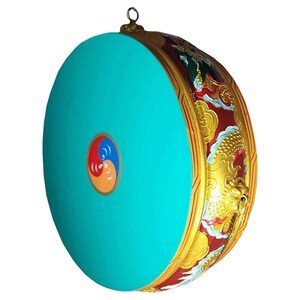 May include: A round, turquoise drum with a gold and red decorative rim. The rim features a dragon design. The drum face has a yin-yang symbol in the center. A small loop is at the top for hanging.