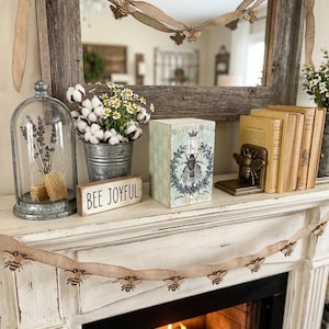Può includere: Una decorazione per mensola del camino con una ghirlanda di api in iuta, una campana di vetro con lavanda e favo, un secchiello in metallo con cotone e margherite, un cartello in legno con la scritta BEE JOYFUL, una scatola e dei libri.