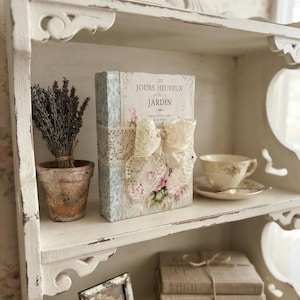 May include: A white distressed wooden shelf holding a decorative book titled Les Jours Heureux du Jardin with a lace bow, a terracotta pot with dried lavender, a porcelain teacup and saucer, a small metal picture frame, and three stacked books.