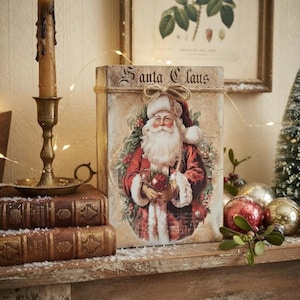 May include: A rectangular wooden box featuring a vintage-style illustration of Santa Claus in a red coat, with the text Santa Claus printed at the top. The box is accented with a jute twine bow and has a distressed, aged finish on its surface.