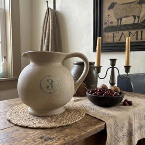 May include: A round, beige ceramic pitcher with a textured surface and a handle on the right side. The front of the pitcher features a circular emblem containing the number 3. The pitcher has a wide, bulbous body and a short, flared spout.