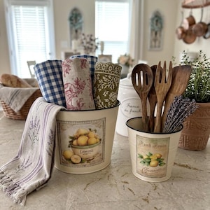 Può includere: Una scena di cucina con due secchi in metallo color crema. Uno contiene canovacci a fantasia arrotolati, l'altro utensili di legno e lavanda. Entrambi i secchi presentano opere d'arte a tema limoni. Un cesto di pane sullo sfondo.