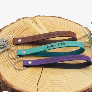 May include: Three leather keychains with different colored straps and engraved text. The top keychain is brown with the text "Your Verse Here". The middle keychain is teal with the text "John 3:16". The bottom keychain is purple with the text "Custom Scripture".