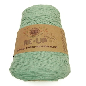May include: A large spool of light green yarn with a brown paper label wrapped around it. The label reads "RE-UP RECYCLED COTTON-POLYESTER BLEND" and features the words "SUSTAINABLE STITCHING" and "Bonus Bundle". The yarn is tightly wound and appears soft.