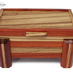 May include: A small wooden box with a hinged lid. The box is made of various shades of wood, including red, brown, and beige. The box has a simple design with a rectangular shape and four legs.