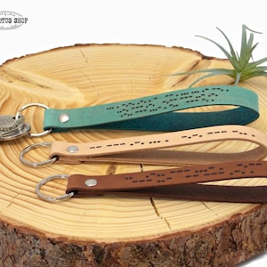 May include: Three leather keychains with different colored leather straps: teal, beige, and brown. Each strap has a metal ring and a pattern of dots embossed on the leather. The keychains are displayed on a wooden surface.