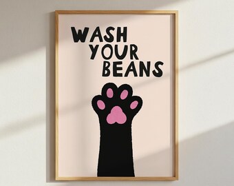 May include: A framed wall art print featuring a light beige background with the black text WASH YOUR BEANS above a graphic illustration of a black animal paw with pink pads. The artwork is enclosed in a simple light-colored wooden frame.