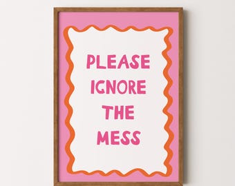 May include: A framed print with the words "PLEASE IGNORE THE MESS" in pink letters. The text is centered on a white background, framed by a wavy orange border and a pink background. The frame is a warm brown color.